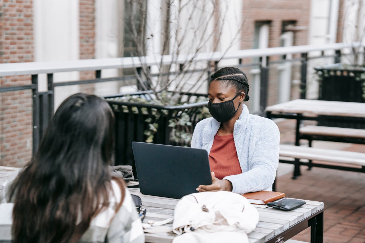 Two women studying outdoors with laptops, wearing masks, maintaining social distance during COVID-19.